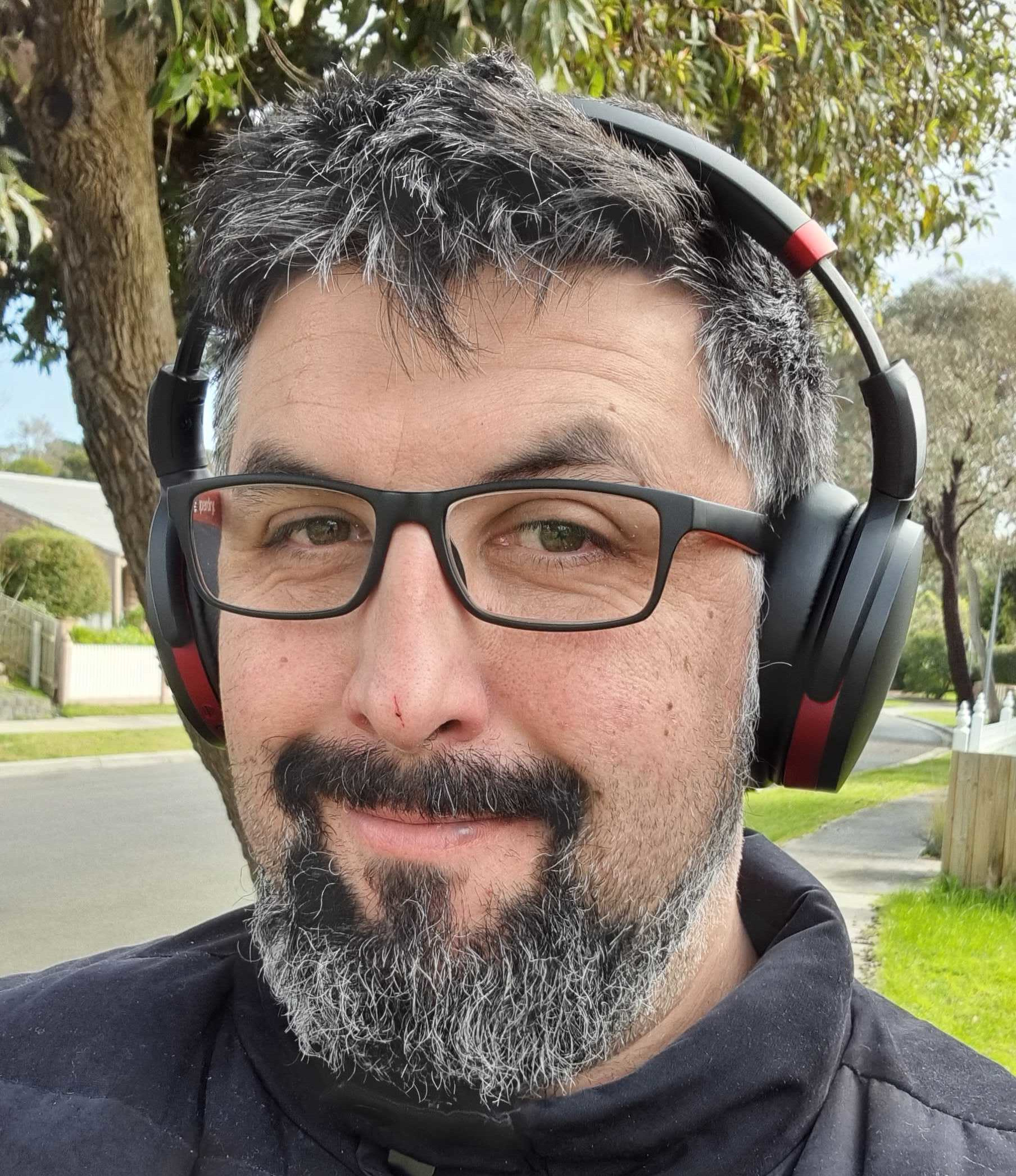 A photograph of Rob Norris, wearing glasses and headphones, out for a walk on a pleasant August day.